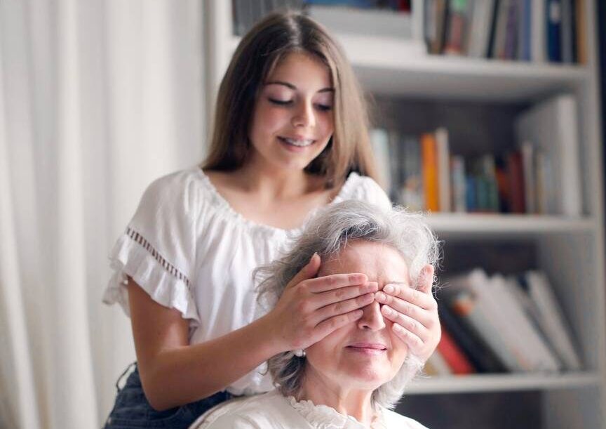 cheerful teenager playing with grandmother guess who game while making surprise in light living room